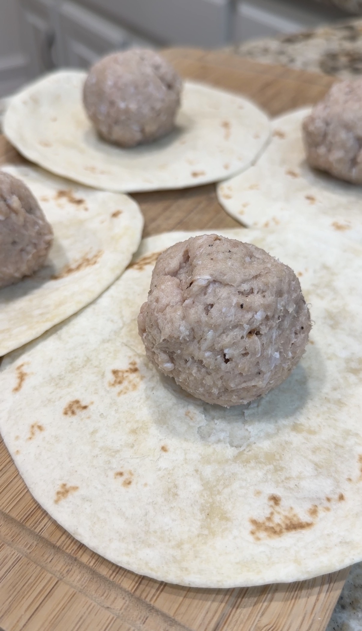 Four uncooked meatballs sit on separate flour tortillas on a wooden surface, ready for preparation. The background shows a kitchen setting out of focus.