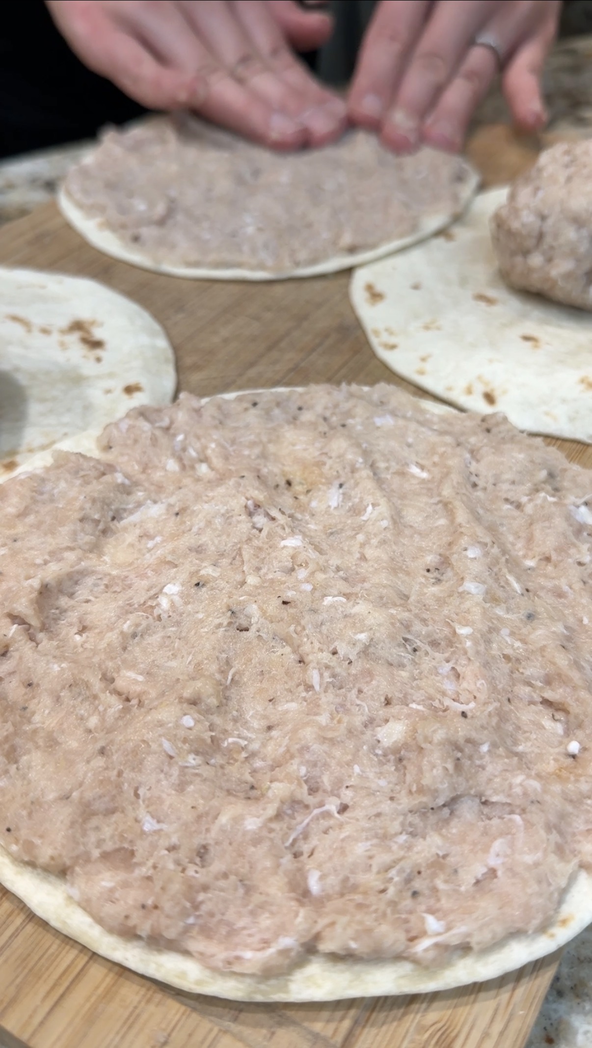 A close-up of raw ground meat spread evenly on a round tortilla with more tortillas and meat in the background. Hands are visible shaping the mixture on a wooden surface.