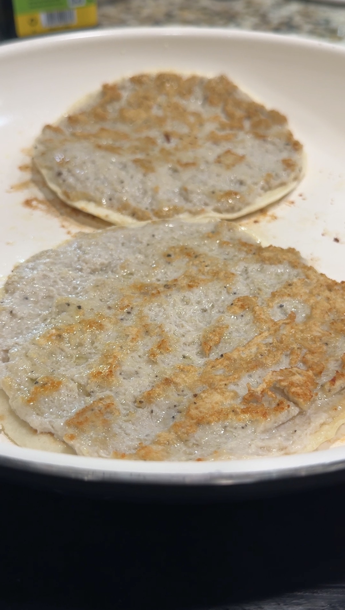Two flat, round smash tacos with a slightly crispy, golden-brown surface are cooking in a white skillet.