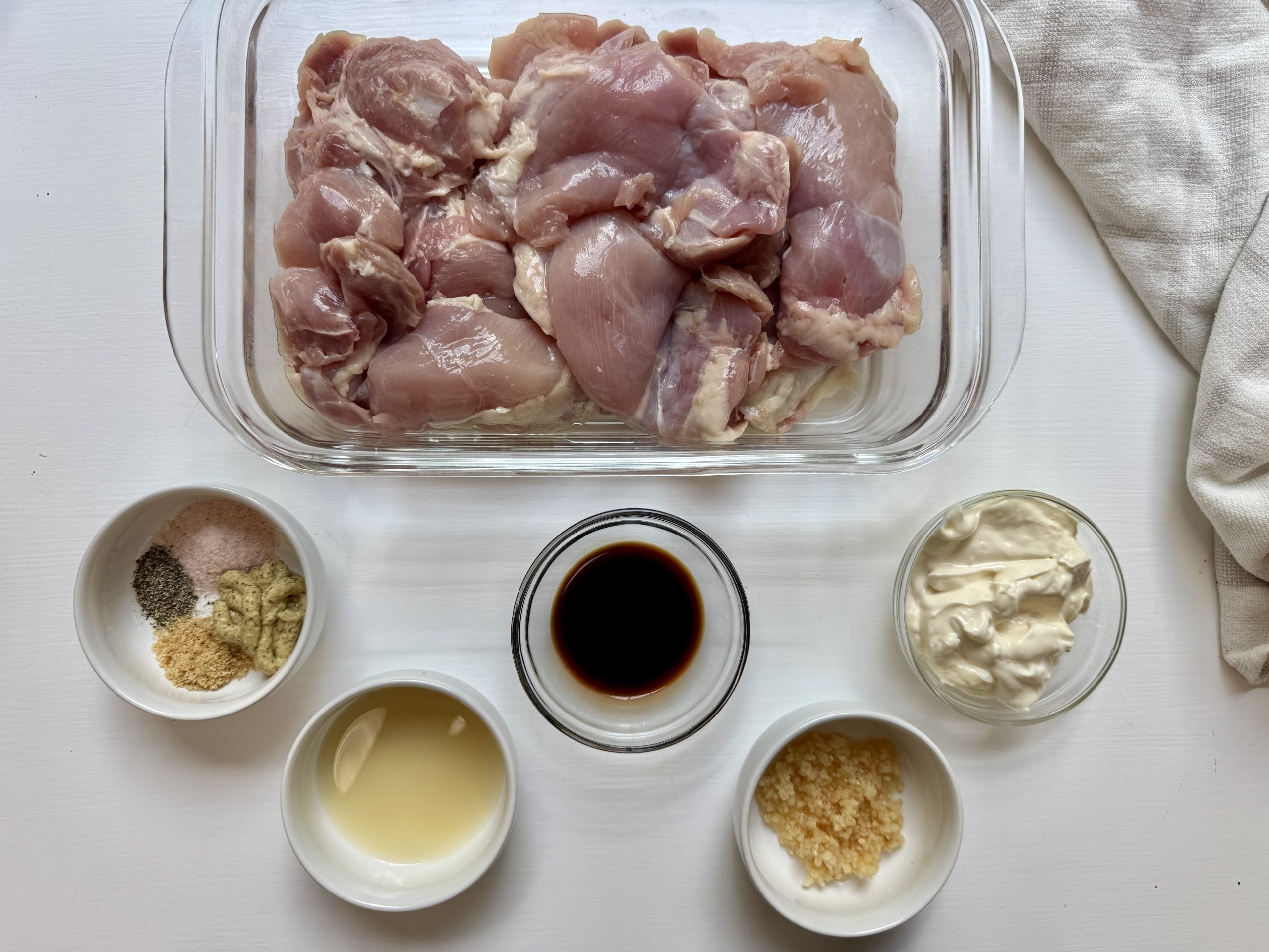 Raw chicken thighs in a glass dish, surrounded by small bowls containing seasonings, minced garlic, mayonnaise, soy sauce, and a liquid ingredient, all arranged on a white surface.