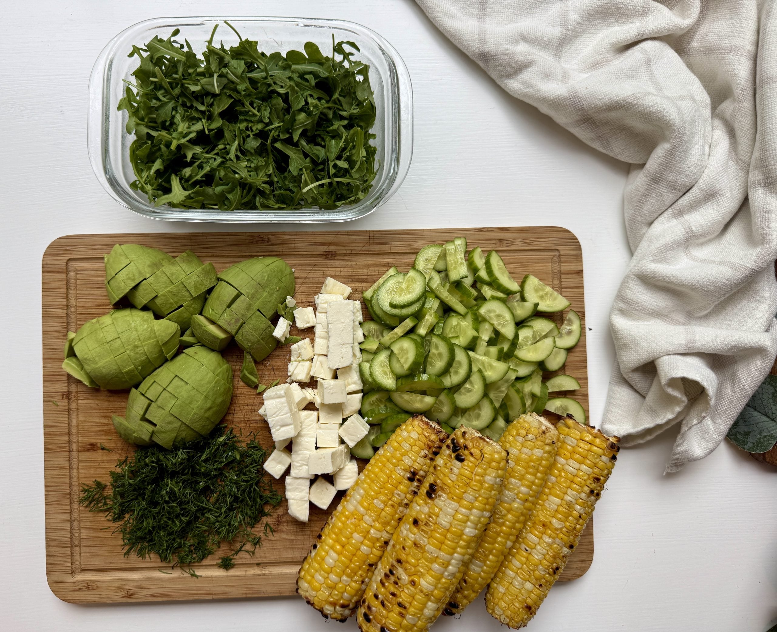 A wooden cutting board holds sliced avocado, chopped cucumber, cubed feta cheese, fresh dill, and grilled corn, with a container of arugula and a white cloth nearby on a white surface.