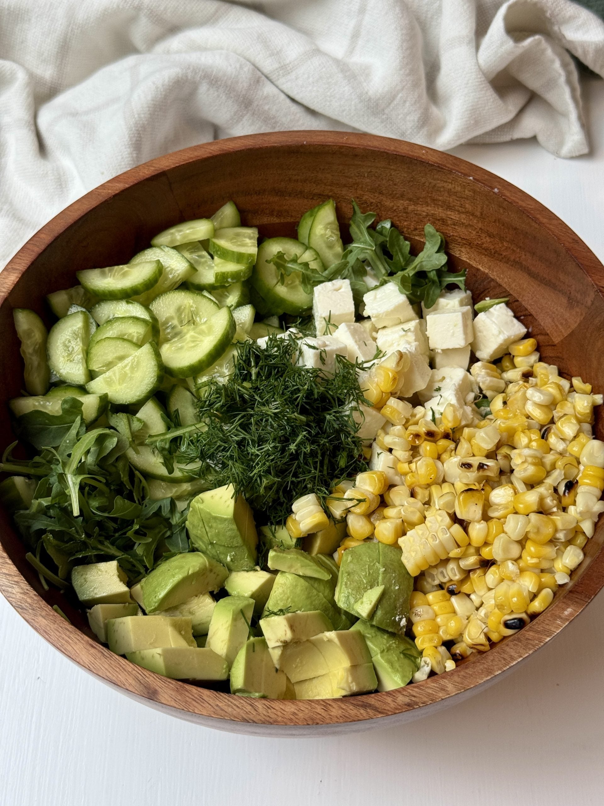 A wooden bowl filled with sliced cucumber, cubed feta cheese, grilled corn kernels, diced avocado, fresh dill, and arugula, arranged in separate sections on a white surface with a cloth in the background.