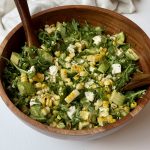 A wooden bowl filled with a fresh salad of arugula, avocado, corn, feta cheese, and fresh herbs, with two wooden salad tongs resting inside the bowl.
