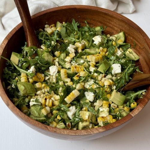 A wooden bowl filled with a fresh salad of arugula, avocado, corn, feta cheese, and fresh herbs, with two wooden salad tongs resting inside the bowl.