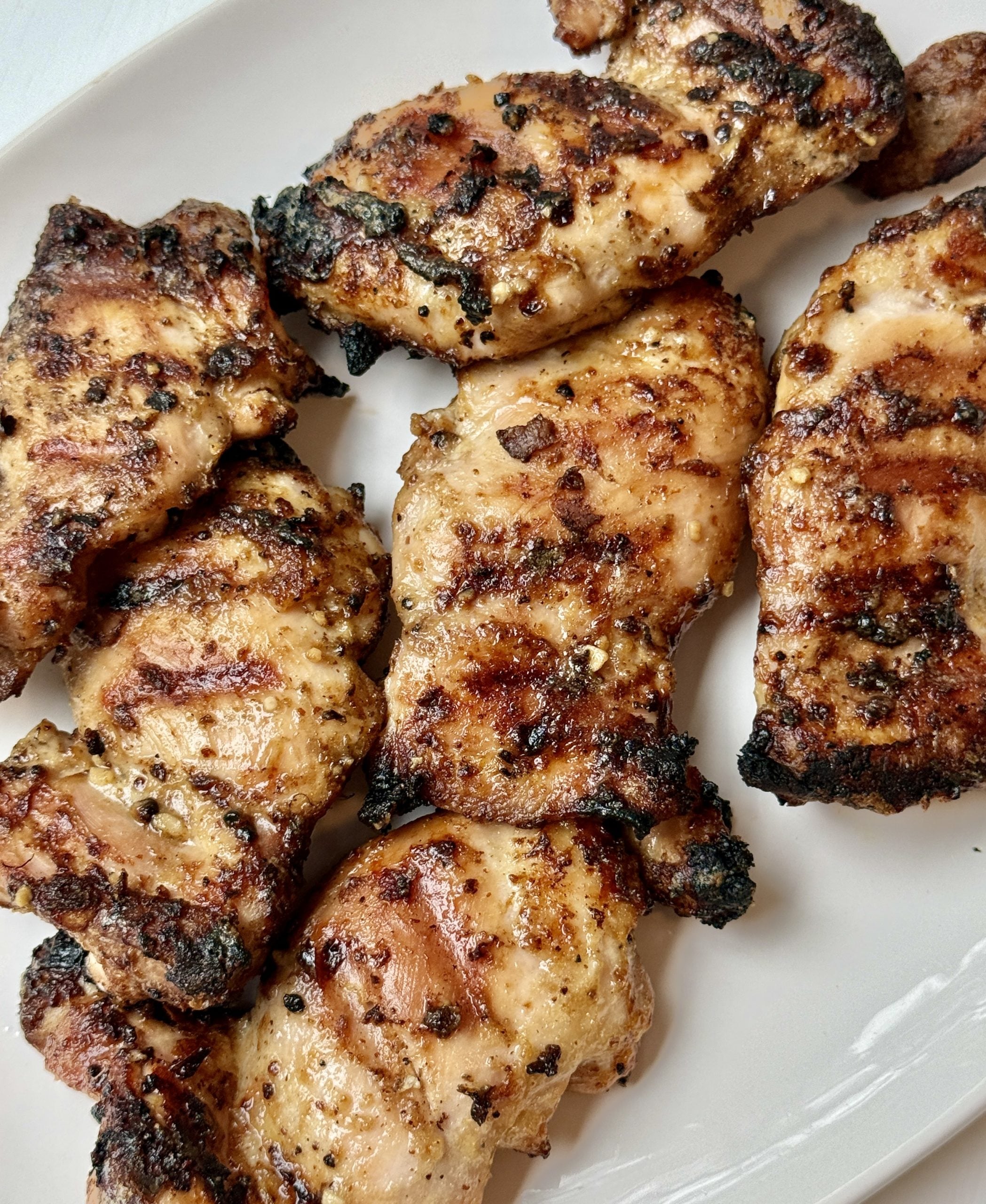 Grilled chicken thighs with visible char marks arranged on a white plate, showing a golden-brown, slightly crispy exterior.