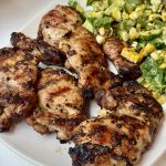 Grilled chicken thighs with visible grill marks served on a white plate alongside a fresh salad with corn, avocado, greens, and herbs.