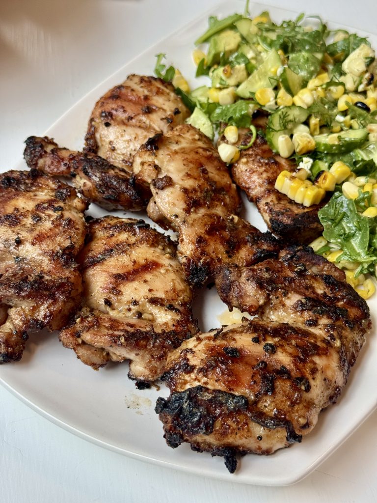 Grilled chicken thighs with visible grill marks served on a white plate alongside a fresh salad with corn, avocado, greens, and herbs.