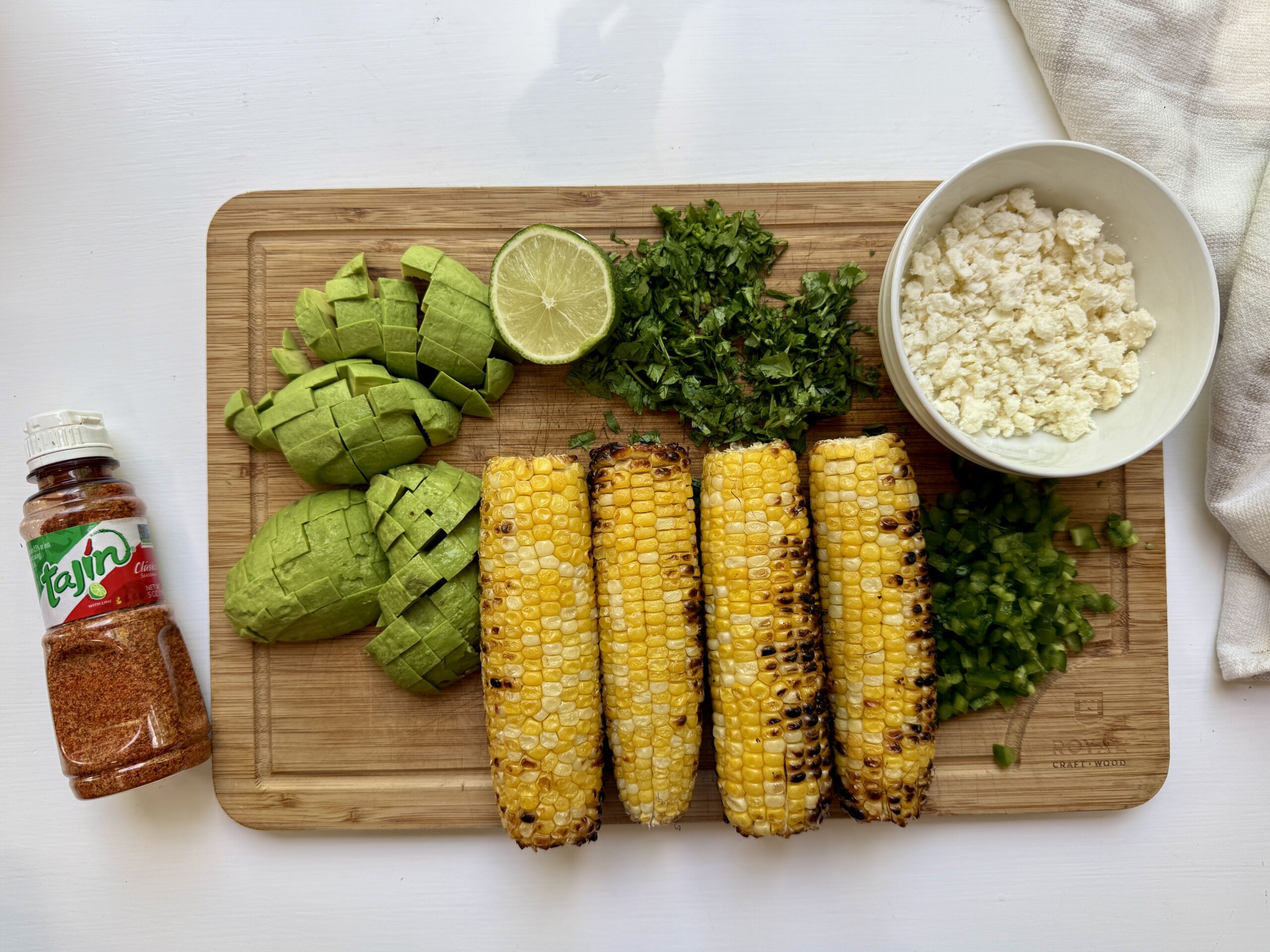 A wooden board holds four grilled corn cobs, chopped avocado, cilantro, jalapeño, half a lime, and a bowl of cheese. A bottle of Tajín seasoning sits beside the board on a white surface.
