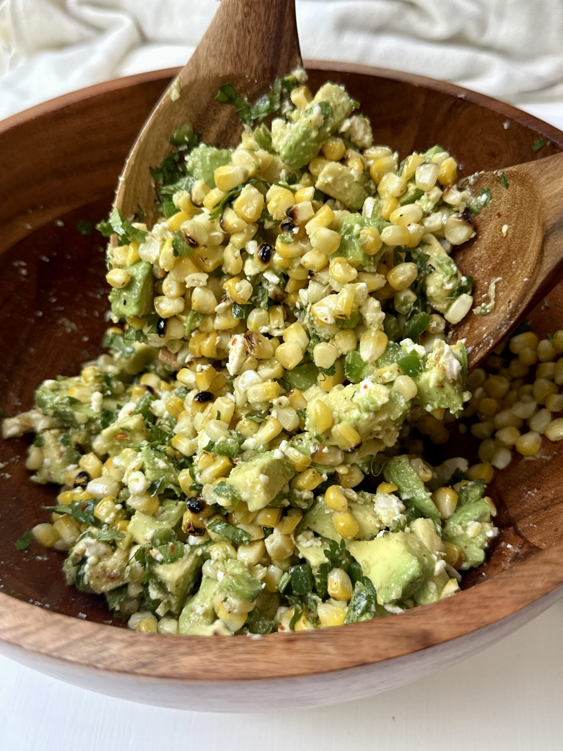 A wooden bowl filled with a salad made of charred corn, diced avocado, chopped cilantro, and cheese, being mixed with two wooden spoons.