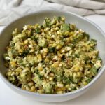 A bowl filled with a salad made of corn, avocado chunks, cheese, and herbs, garnished with spices, placed on a white surface with a light cloth in the background.