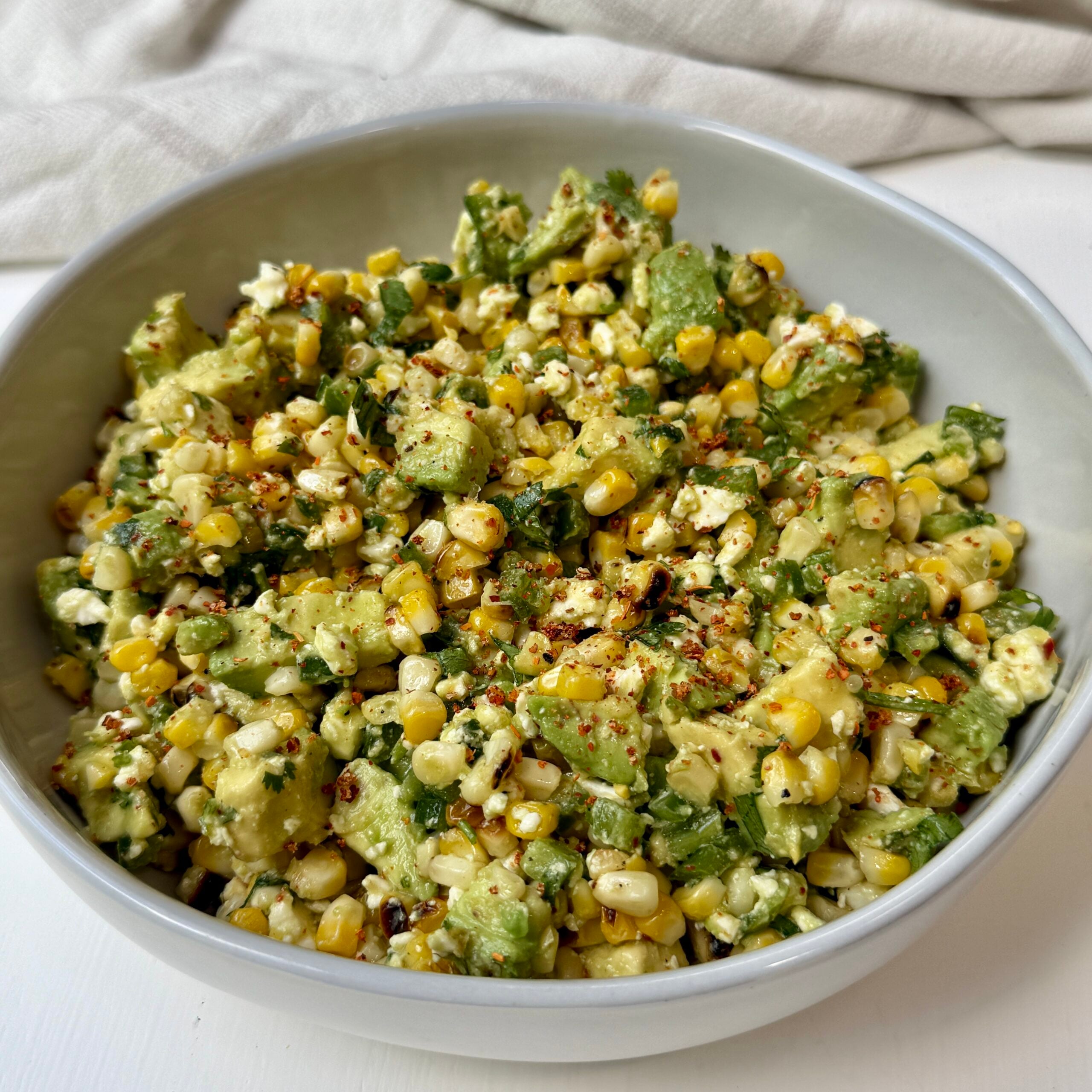 A bowl filled with a salad made of corn, avocado chunks, cheese, and herbs, garnished with spices, placed on a white surface with a light cloth in the background.