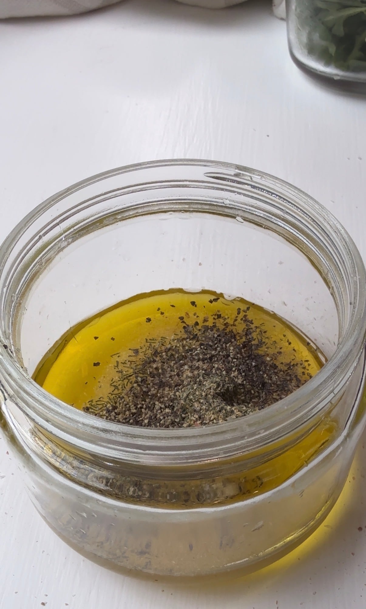 A clear glass jar filled with olive oil, topped with a mound of ground black pepper and dried herbs, sits on a white surface.