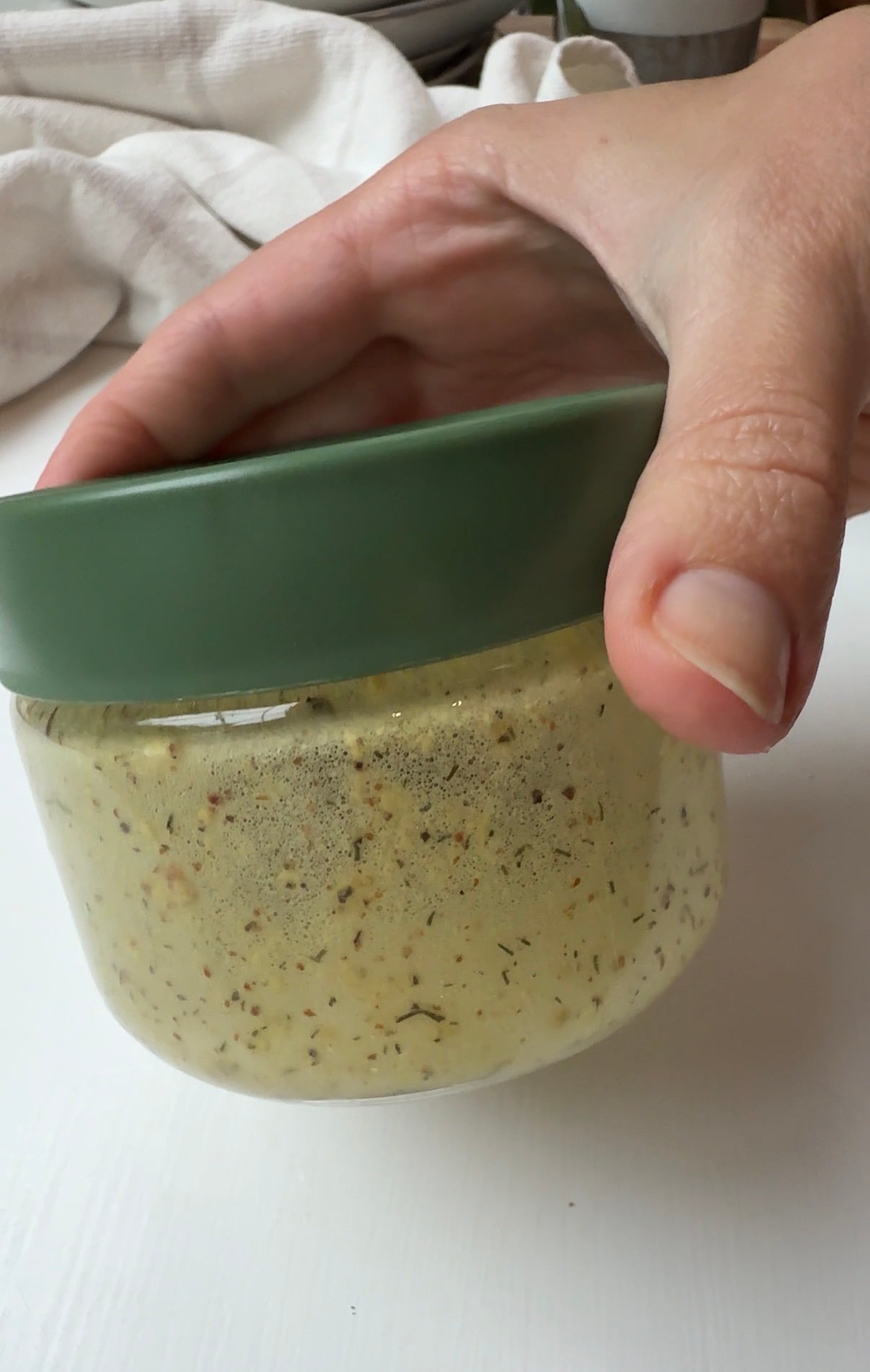A hand holds a glass jar with a green lid, containing a creamy, speckled mixture, possibly a homemade sauce or dressing, against a white background.
