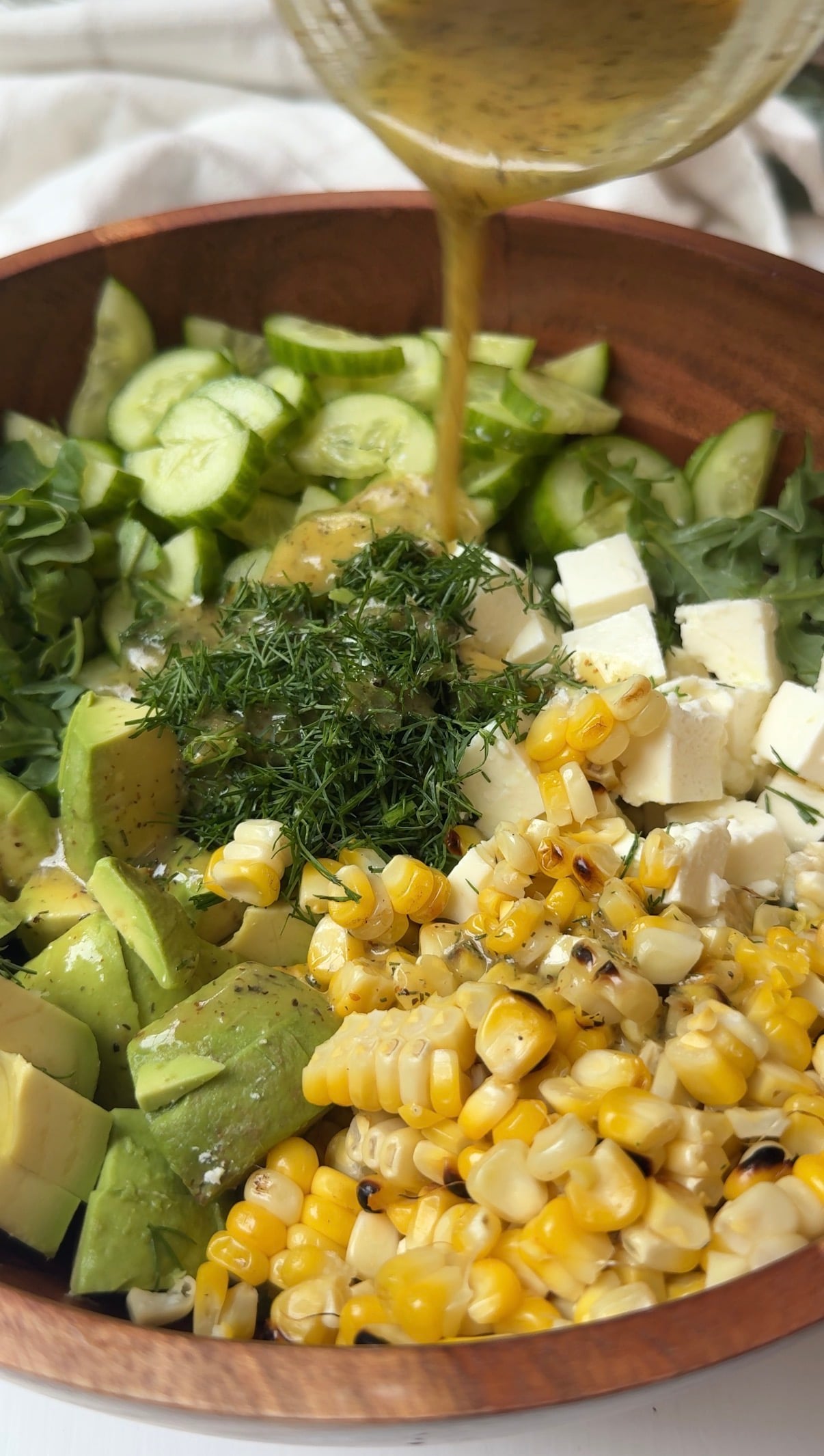 A wooden bowl filled with sliced cucumber, arugula, avocado, grilled corn, feta cheese cubes, and fresh dill, with a dressing being poured over the salad.