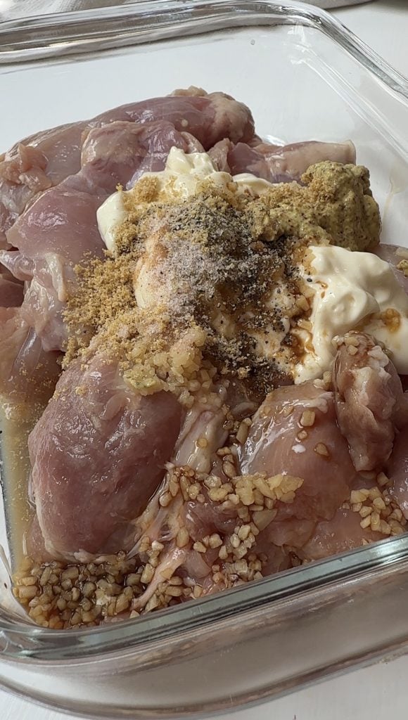 Raw chicken pieces in a glass dish topped with chopped garlic, mayonnaise, mustard, black pepper, and seasoning, ready to be mixed for marination.