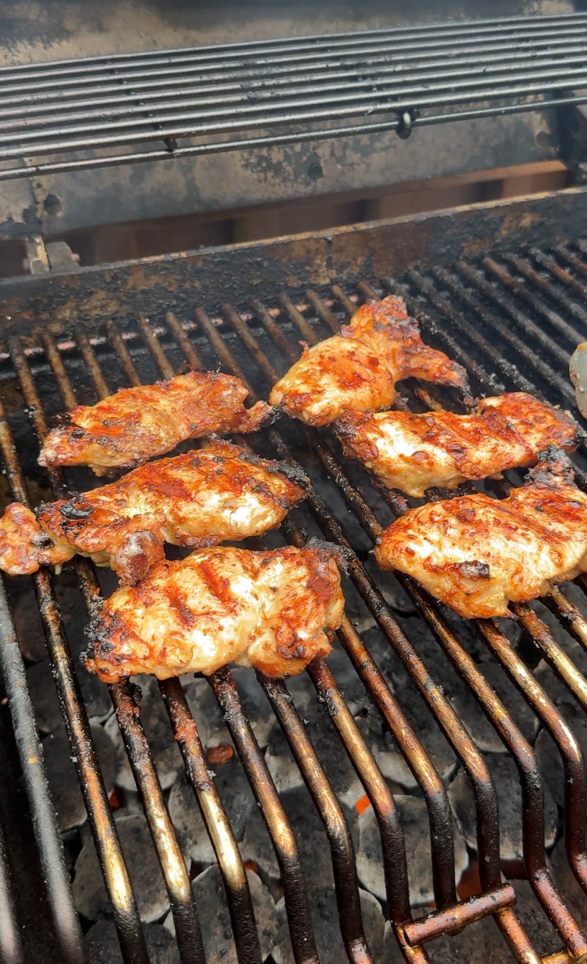Seasoned chicken wings cooking on a barbecue grill, showing grill marks and some charring, with smoke rising and hot coals visible below the grate.