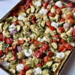 A sheet pan filled with roasted vegetables, chicken pieces, cherry tomatoes, red onions, zucchini, red bell peppers, dollops of white cheese, and drizzles of green pesto sauce on parchment paper.