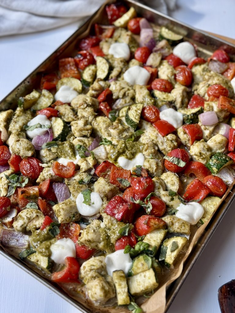 A sheet pan filled with roasted vegetables, chicken pieces, cherry tomatoes, red onions, zucchini, red bell peppers, dollops of white cheese, and drizzles of green pesto sauce on parchment paper.
