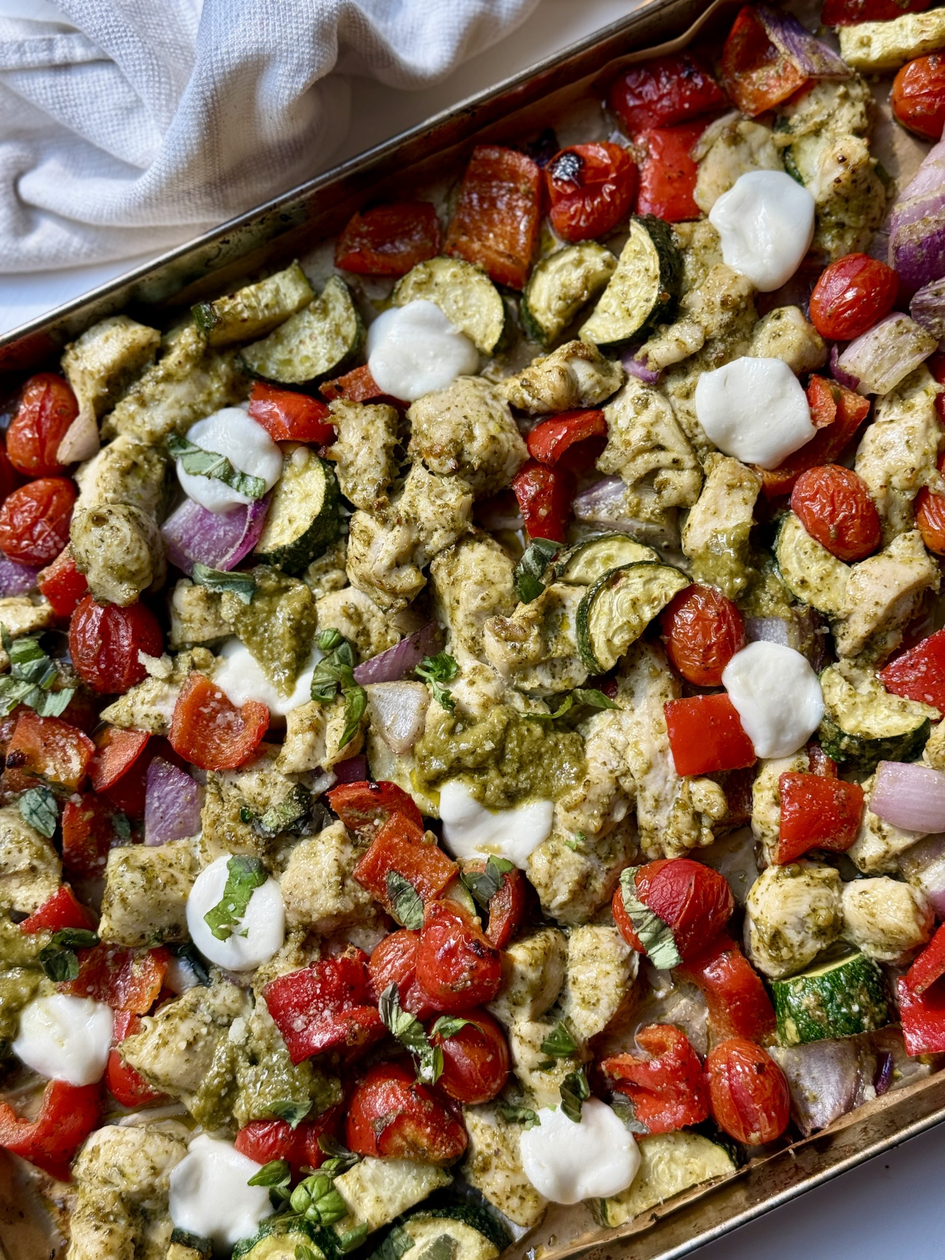 A sheet pan filled with roasted chicken, zucchini, cherry tomatoes, red bell pepper, and red onion, topped with dollops of melted mozzarella cheese and drizzled with green pesto. A white cloth is partly visible in the corner.