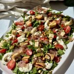 A fresh salad on a white platter with arugula, cherry tomatoes, mozzarella, croutons, prosciutto, pesto drizzle, and crushed pistachios, set on a white table with a light cloth in the background.