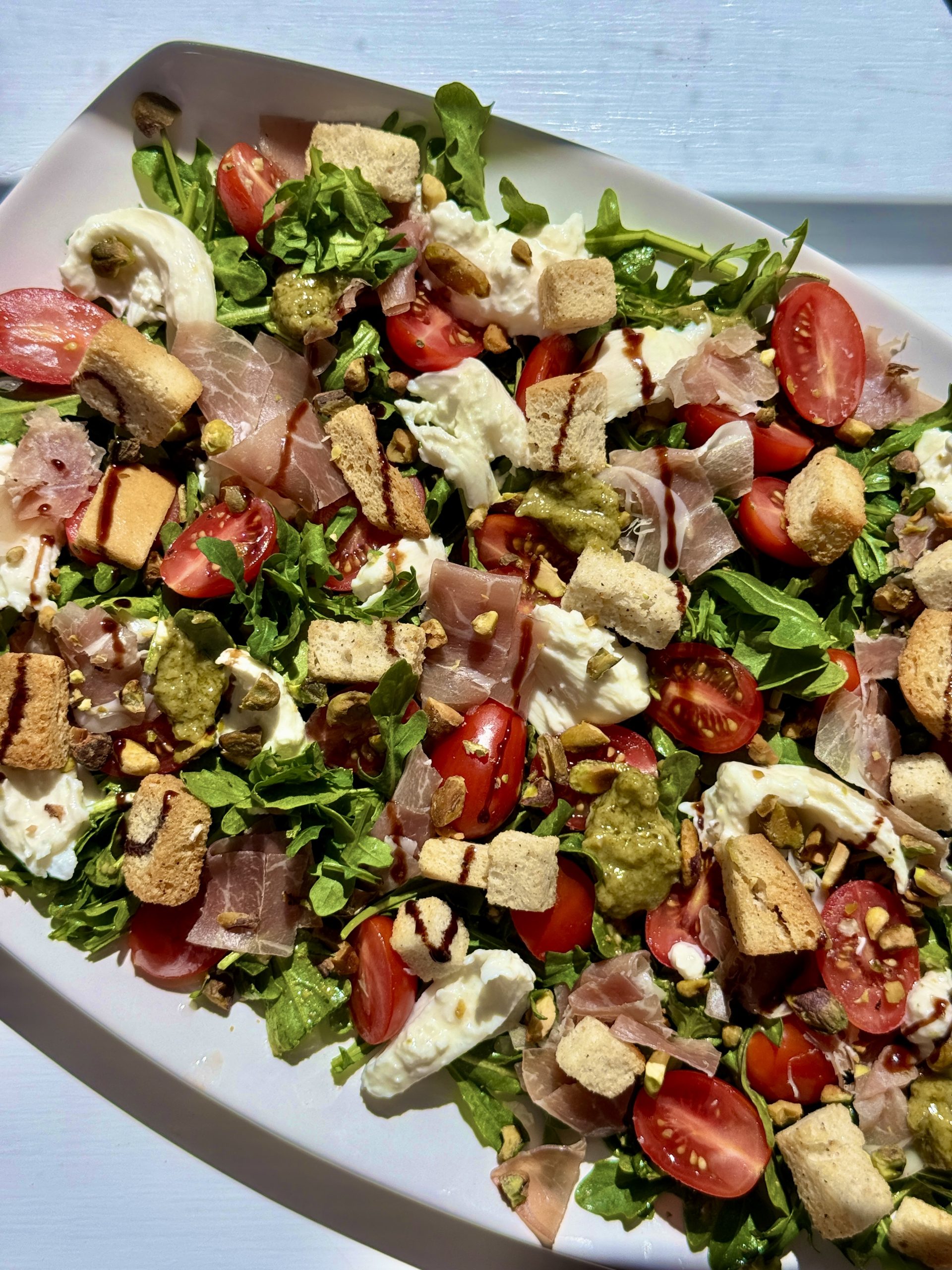 A fresh salad with arugula, mozzarella, cherry tomatoes, prosciutto, croutons, pesto, balsamic glaze, and seeds served on a white plate.