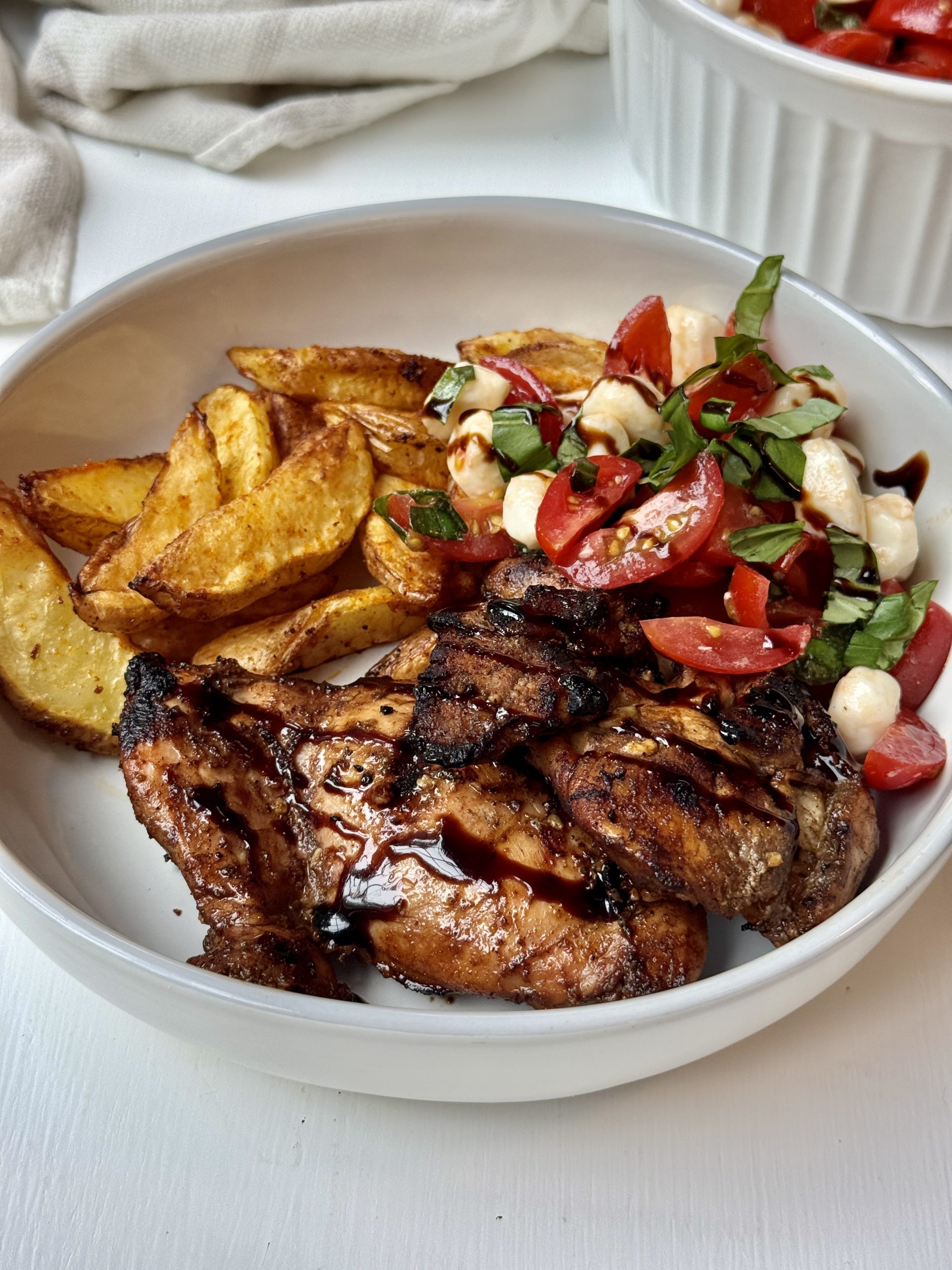 A white bowl with grilled chicken, roasted potato wedges, and a tomato, mozzarella, and basil salad drizzled with balsamic glaze. A bowl of extra salad is in the background on a white surface.