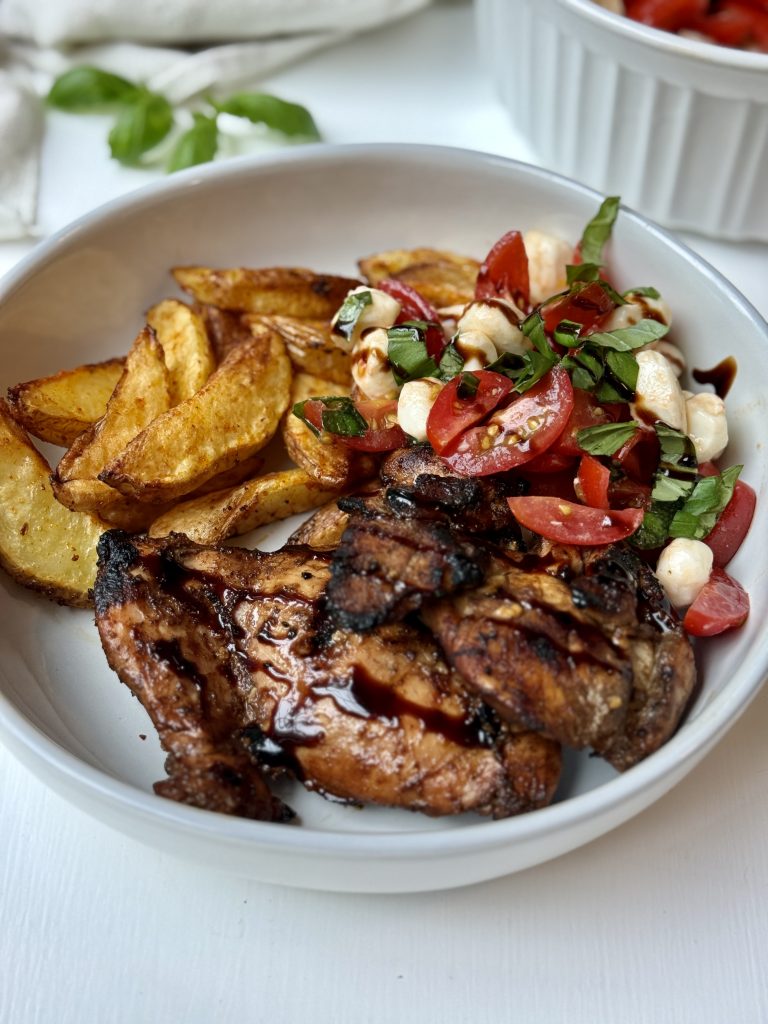 A white bowl with grilled chicken, roasted potato wedges, and a tomato, mozzarella, and basil salad drizzled with balsamic glaze. A bowl of extra salad is in the background on a white surface.
