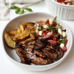 A white bowl with grilled chicken, roasted potato wedges, and a tomato, mozzarella, and basil salad drizzled with balsamic glaze. A bowl of extra salad is in the background on a white surface.