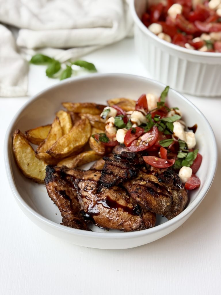 A white bowl with grilled chicken, roasted potato wedges, and a tomato, mozzarella, and basil salad drizzled with balsamic glaze. A bowl of extra salad is in the background on a white surface.