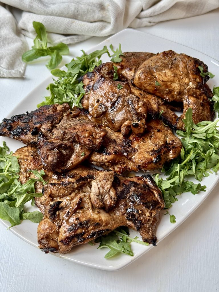 Grilled, seasoned pieces of chicken laid on a bed of fresh green arugula on a white rectangular plate, with a white cloth in the background.