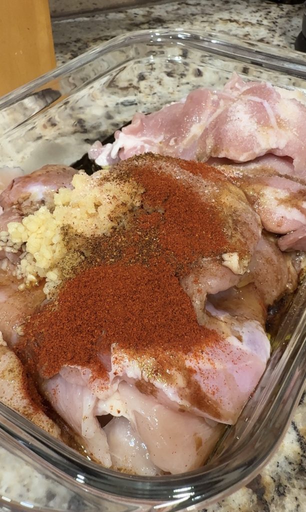 Raw chicken pieces in a glass baking dish topped with minced garlic, paprika, black pepper, and other spices, set on a speckled kitchen countertop.