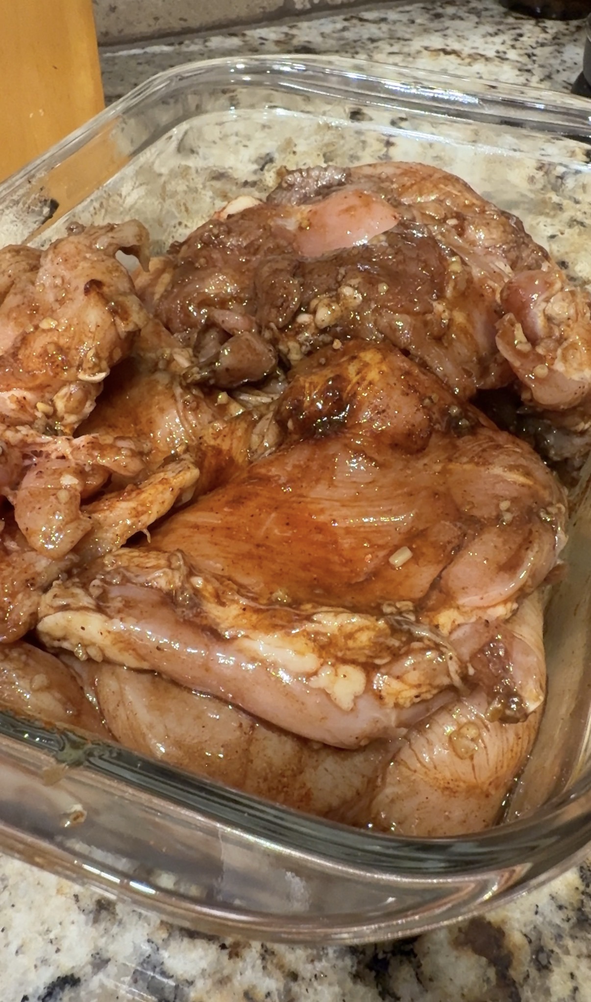 Raw chicken pieces marinated in a reddish-brown spice mixture are arranged in a glass dish on a granite countertop, ready for cooking.