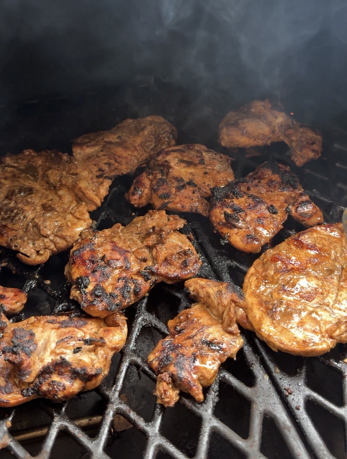 Marinated pieces of meat are grilling on a barbecue, with visible char marks and smoke rising from the hot grill grates.