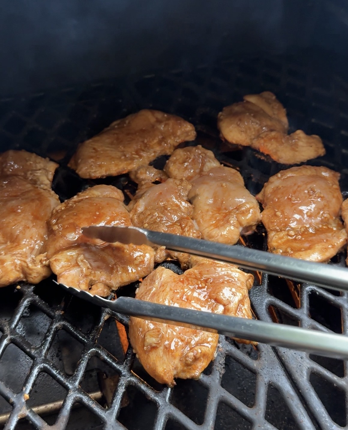Marinated pieces of meat being grilled on a barbecue with metal tongs holding one piece over a black, diamond-patterned grill grate.