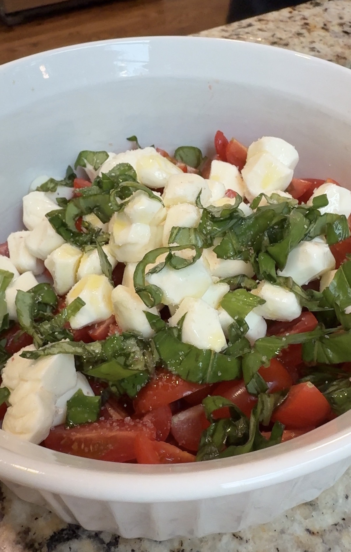 A white bowl filled with a salad of chopped tomatoes, mozzarella cubes, and fresh basil ribbons, drizzled with olive oil, on a granite countertop.