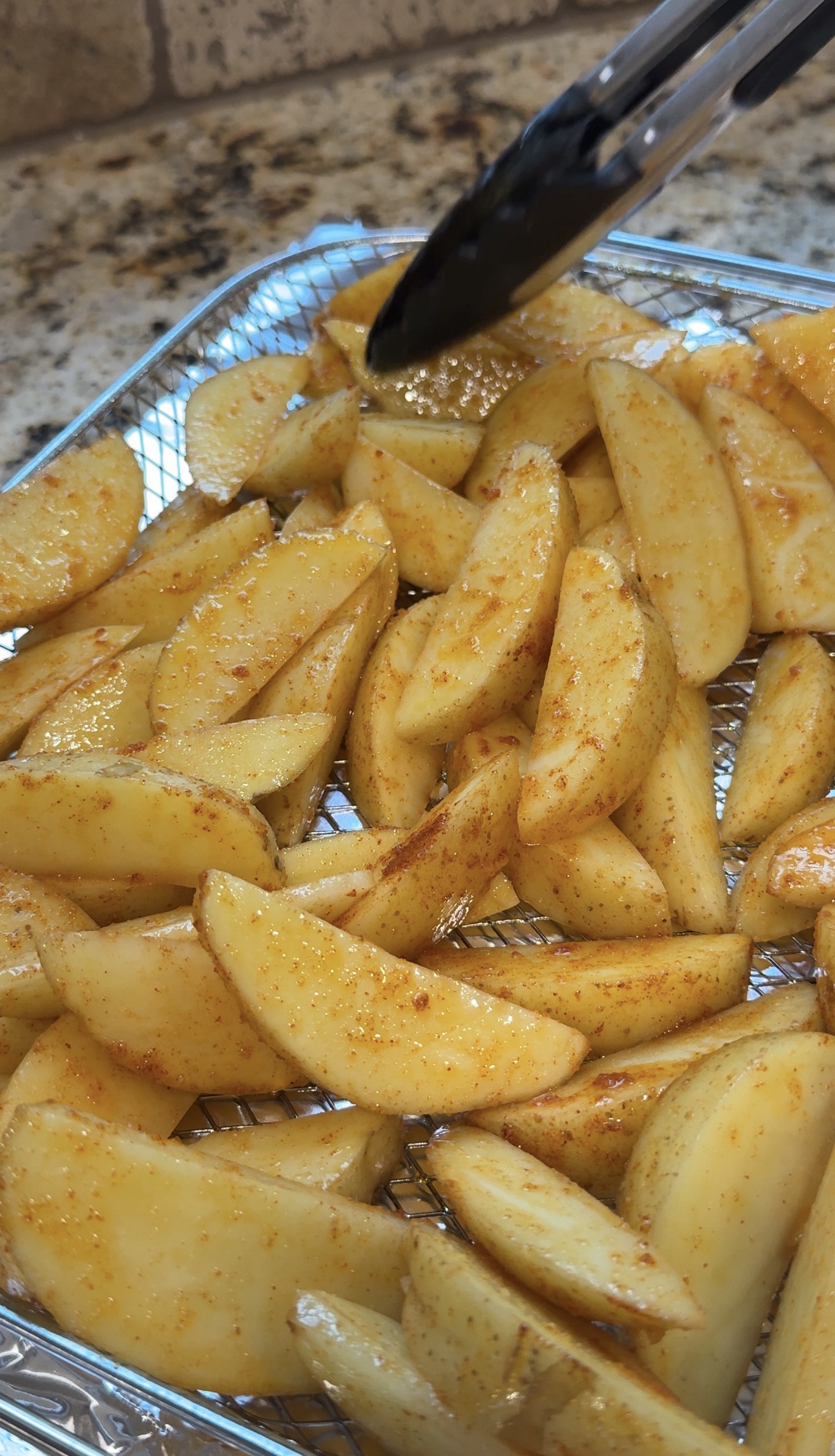Seasoned potato wedges on a wire rack, ready for cooking, with a pair of black tongs holding one wedge. The potatoes are coated with spices and arranged on a kitchen countertop.