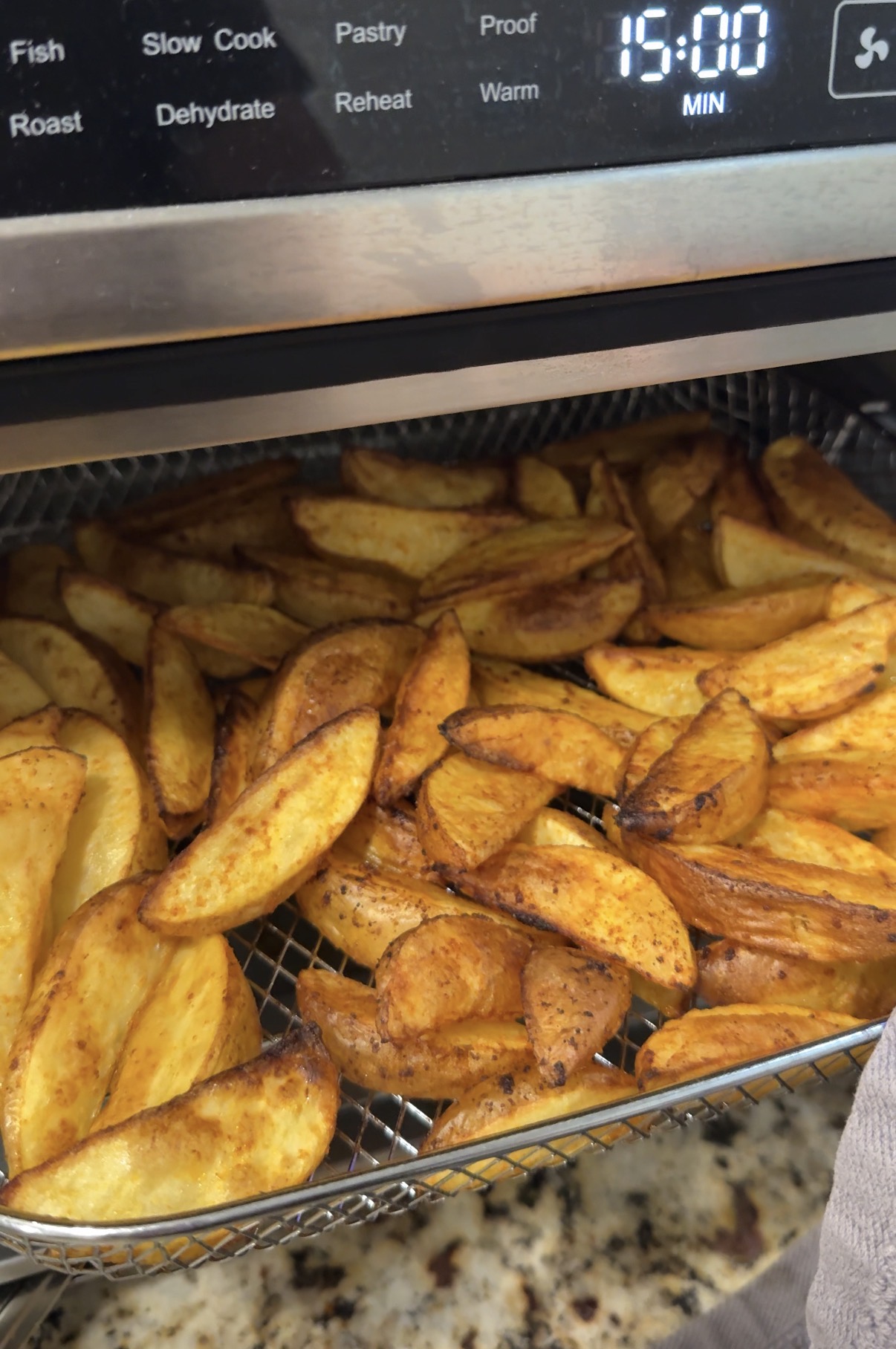 Air fryer basket filled with golden, seasoned potato wedges beneath a digital display reading 15:00 minutes. The basket sits on a granite countertop.