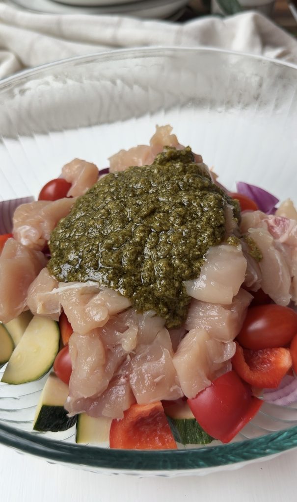 A glass bowl filled with raw diced chicken, chopped vegetables including zucchini, cherry tomatoes, red onion, and red bell pepper, topped with a generous scoop of green pesto sauce.
