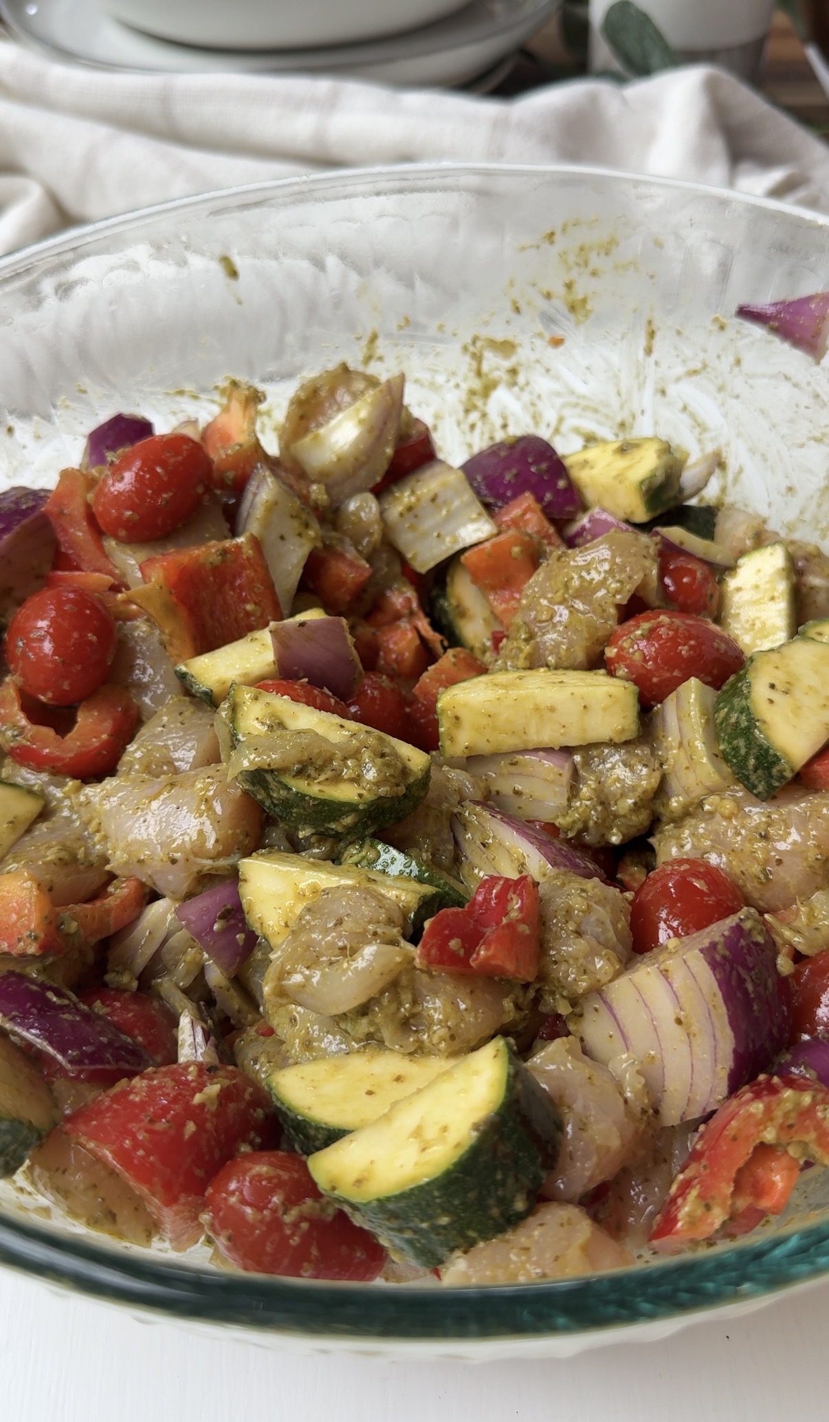 A close-up of a bowl filled with chopped vegetables, including zucchini, red onion, cherry tomatoes, and red bell pepper, all coated in a green herb marinade.