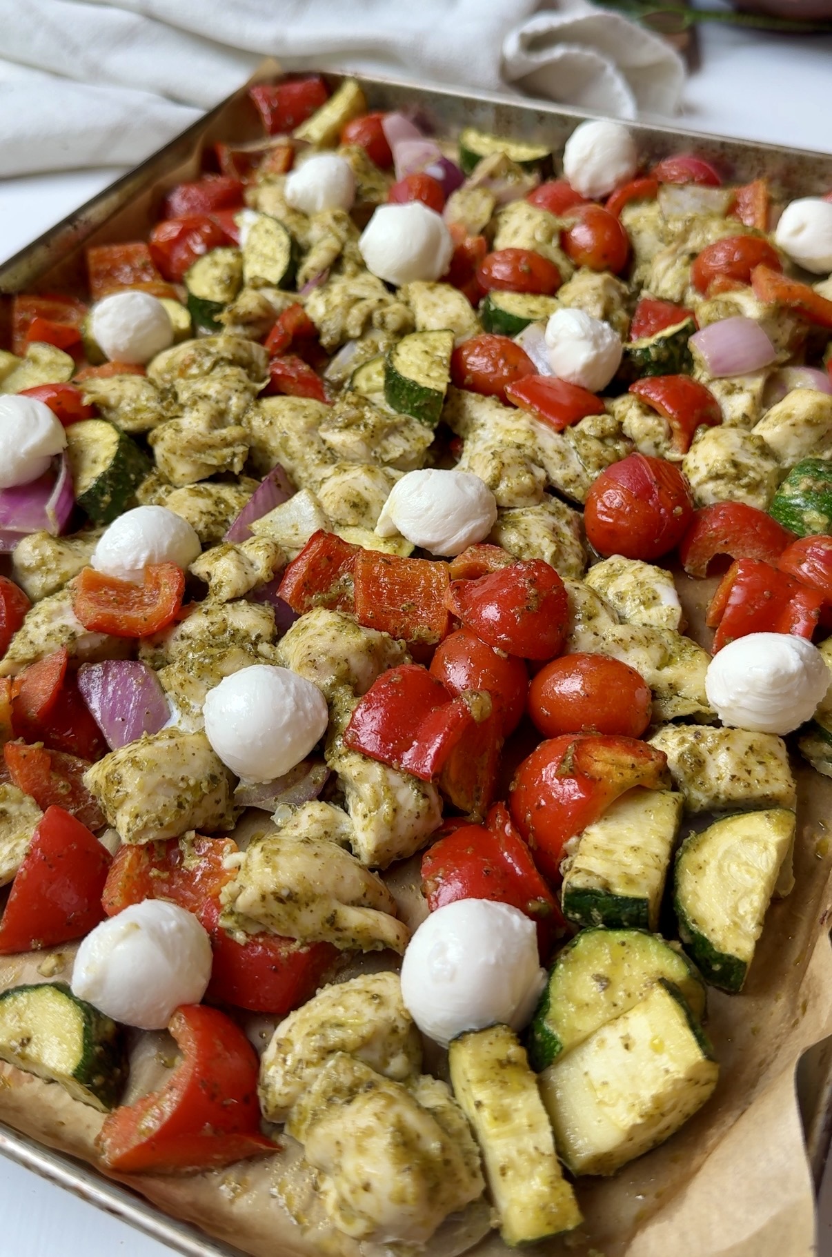 A sheet pan filled with chopped chicken, zucchini, red bell peppers, red onions, cherry tomatoes, mozzarella balls, and a green pesto sauce, all ready to be baked.