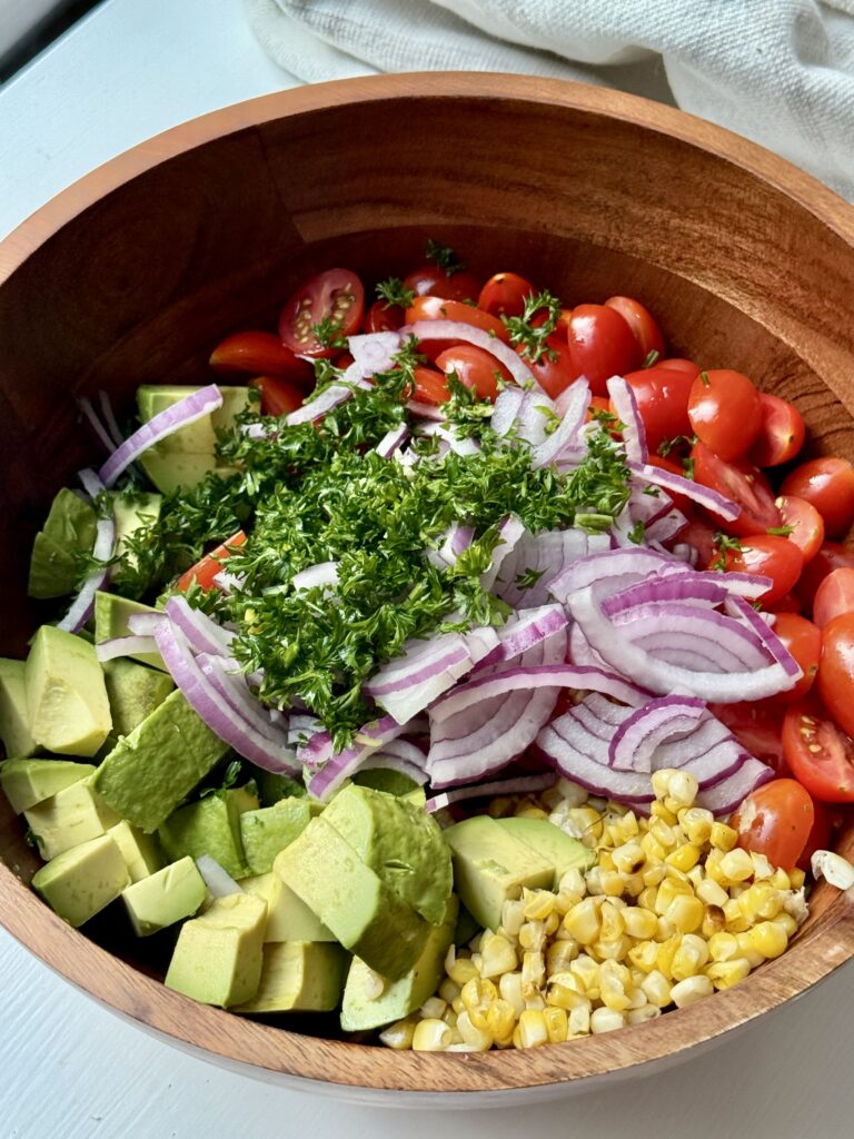 A wooden bowl filled with halved cherry tomatoes, sliced red onions, diced avocado, chopped parsley, and corn kernels, ready to be mixed for a fresh salad.