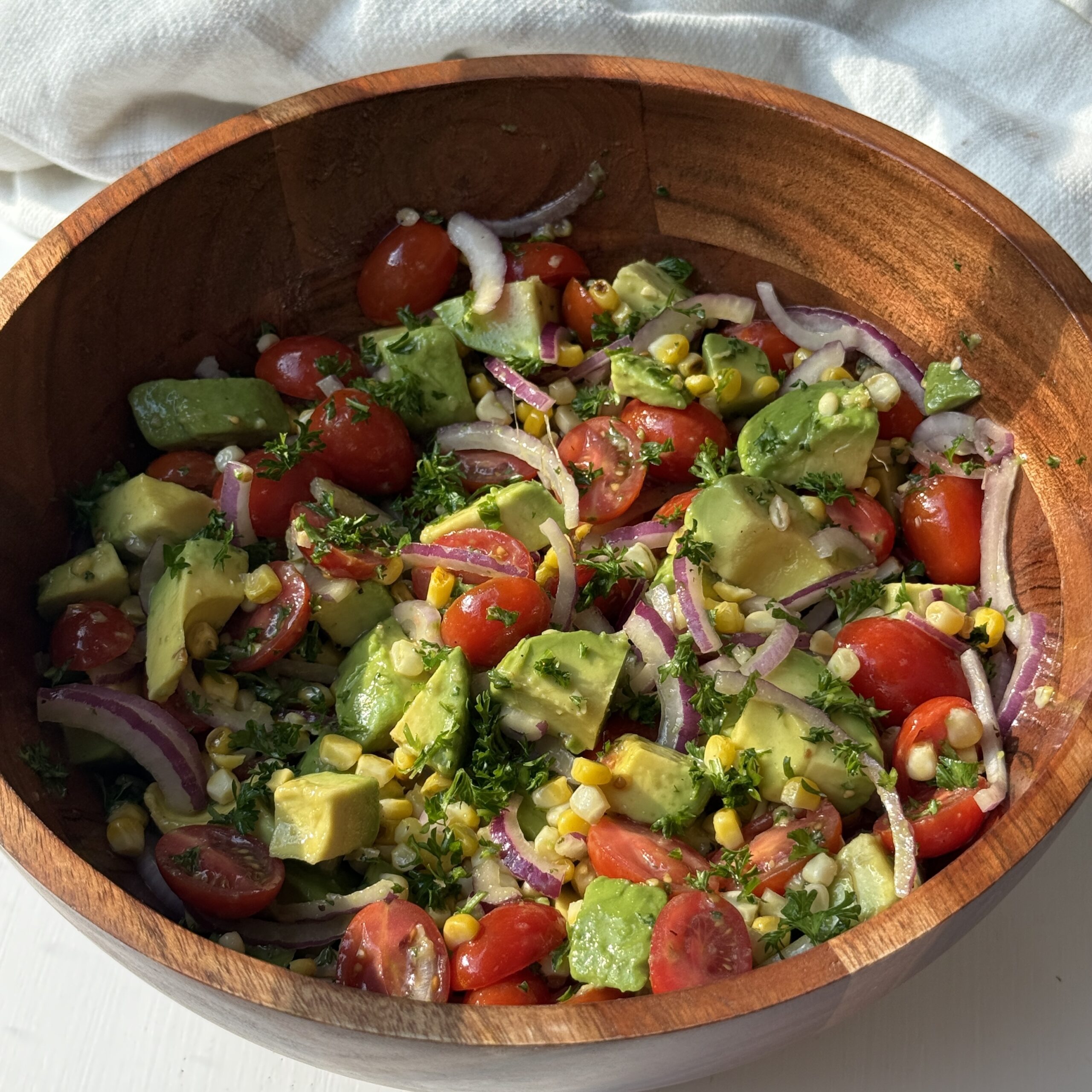 A wooden bowl filled with a fresh salad made of cherry tomatoes, avocado, corn, red onion slices, and herbs, all tossed together and sitting on a white surface with a white cloth in the background.