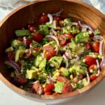 A wooden bowl filled with a fresh salad made of cherry tomatoes, avocado, corn, red onion slices, and herbs, all tossed together and sitting on a white surface with a white cloth in the background.
