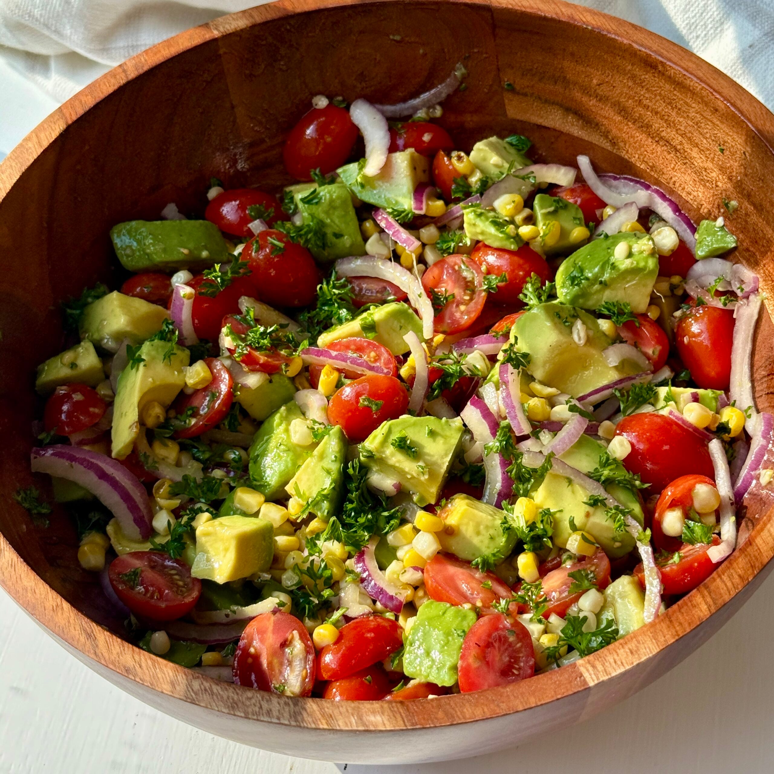 A wooden bowl filled with a fresh salad made of cherry tomatoes, avocado, corn, red onion slices, and herbs, all tossed together and sitting on a white surface with a white cloth in the background.