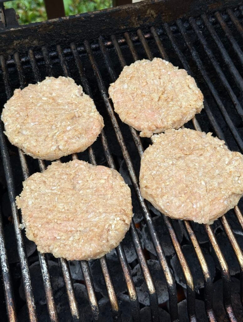 Four raw hamburger patties are cooking on a black grill with grates, outdoors. The patties are uncooked and light in color, with grill marks starting to form underneath.
