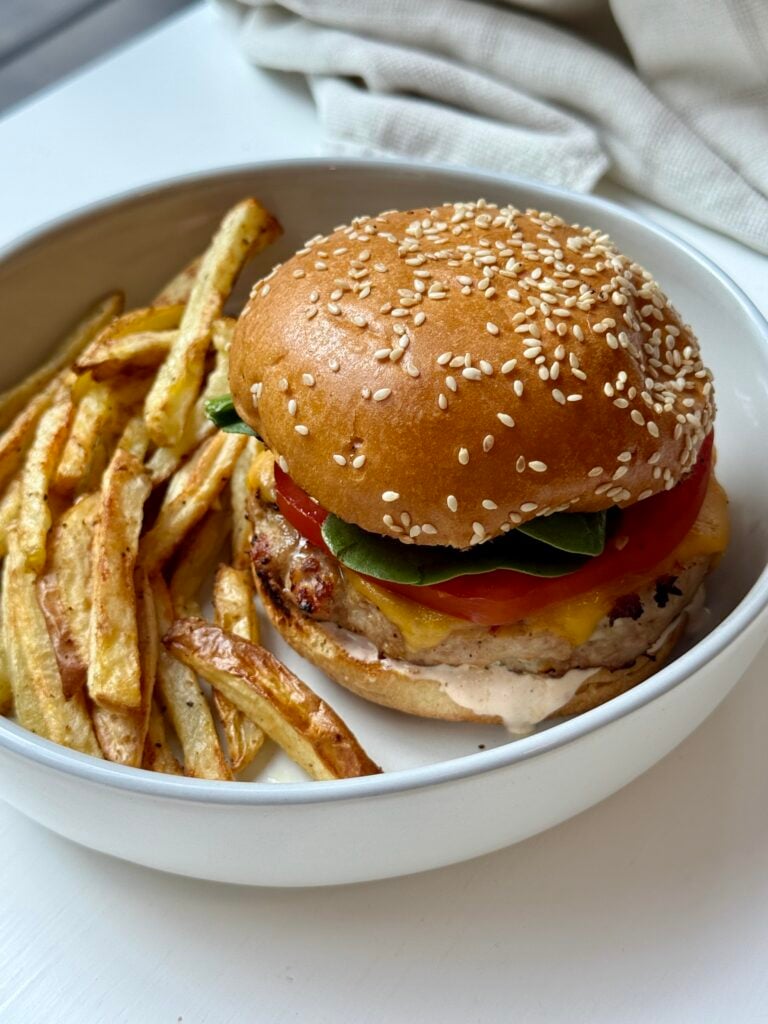 A sesame seed bun chicken burger with tomato, spinach, and cheese served on a white plate with a side of golden French fries.
