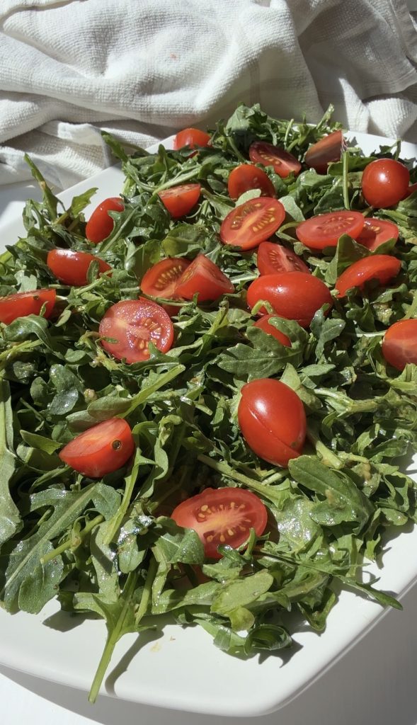 A fresh arugula salad topped with halved cherry tomatoes, lightly dressed and served on a white plate, with a white cloth in the background.