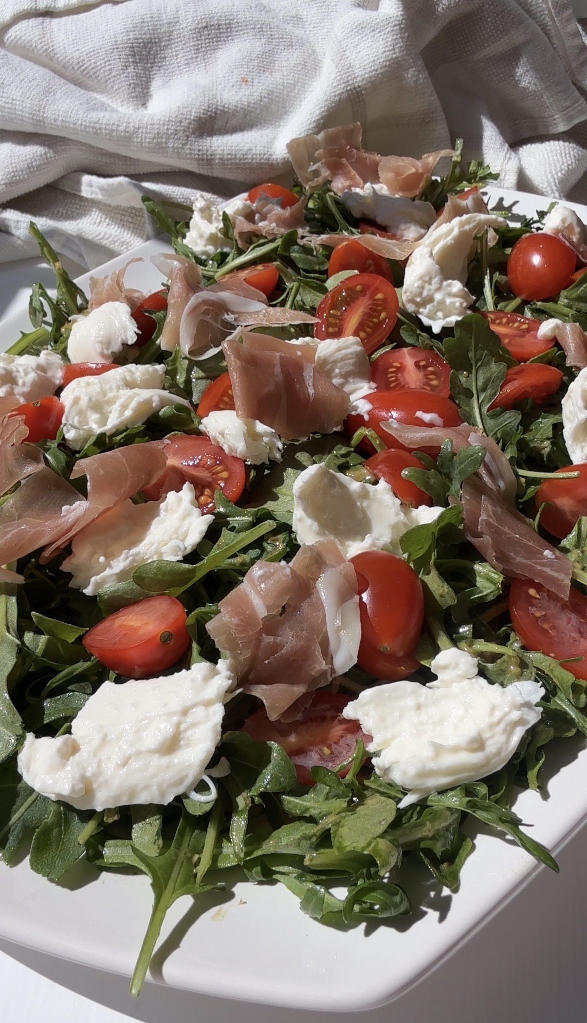 A fresh salad with arugula, cherry tomatoes, slices of prosciutto, and dollops of mozzarella cheese, served on a white plate with a white cloth in the background.