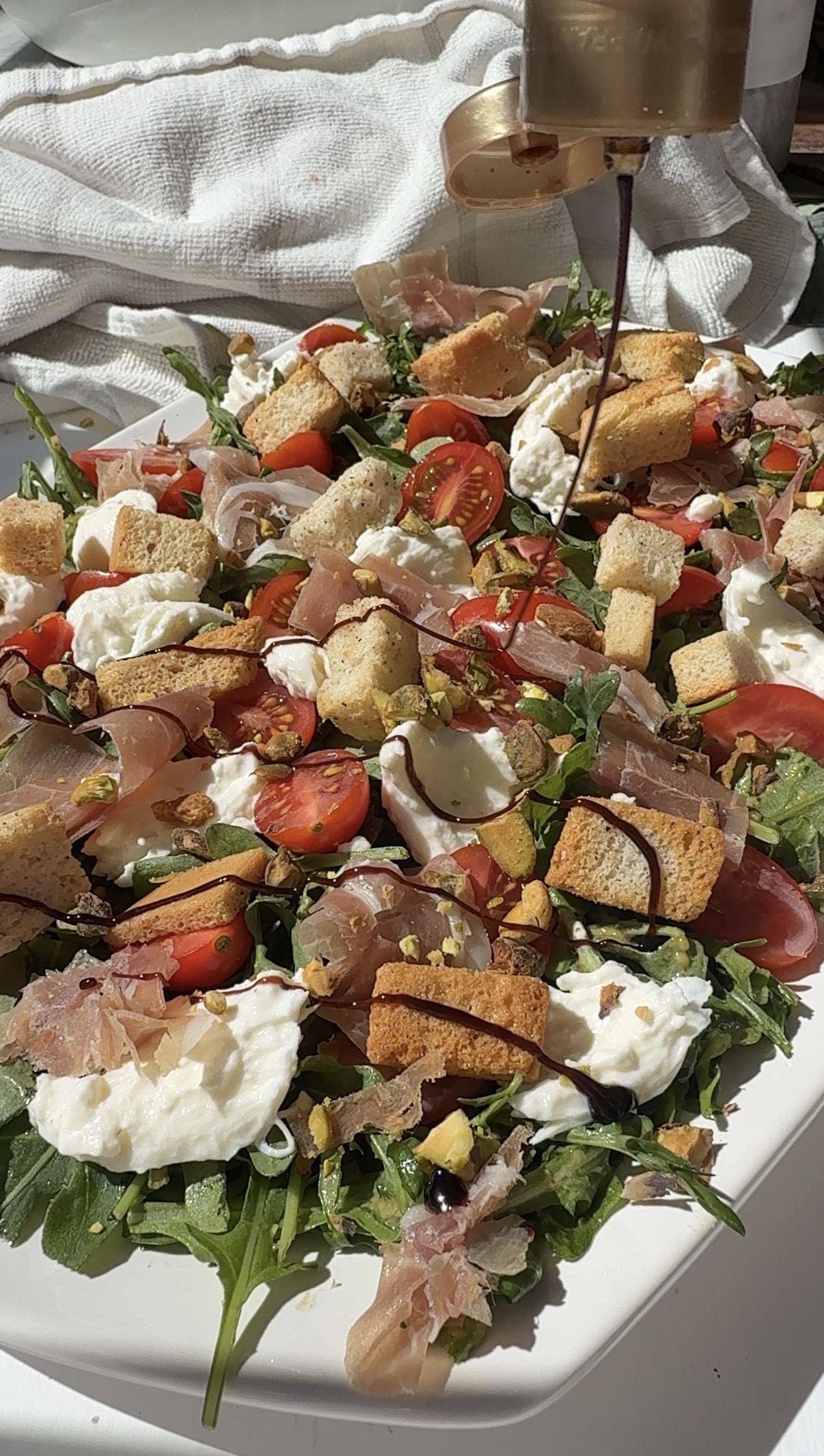 A fresh salad with arugula, cherry tomatoes, burrata cheese, prosciutto, croutons, and chopped pistachios is being drizzled with balsamic glaze on a white platter in bright sunlight.