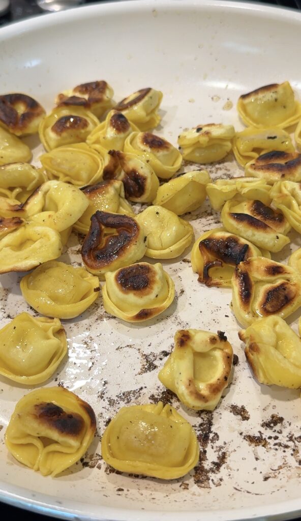 Pan-fried tortellini with golden brown and slightly burnt spots on a white skillet, showing a mix of crispy and soft pasta pieces.